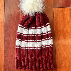 May include: A knitted winter hat in burgundy with white horizontal stripes. The hat features a large, fluffy, cream-colored pom-pom on top. The hat is made of a soft, warm material, perfect for cold weather.
