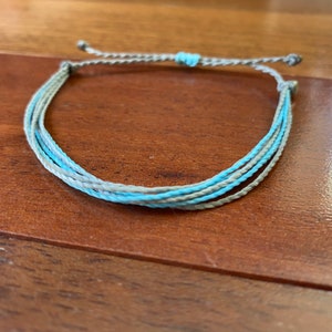 Puede incluir: Una pulsera hecha a mano con múltiples hebras de cordón azul claro y gris. La pulsera tiene un cierre ajustable con una pequeña cuenta de acento azul claro. La pulsera se muestra sobre una superficie de madera.