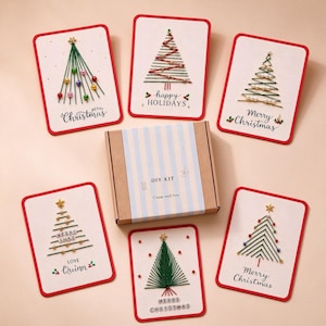 May include: A DIY kit box with blue and white stripes labeled "DIY KIT Create with love" surrounded by six white greeting cards featuring embroidered Christmas tree designs with colorful beads, stars, and text including "Merry Christmas" and "happy HOLIDAYS".