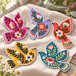 May include: Five embroidered bird-shaped patches featuring intricate floral and leaf patterns. The birds are colored in vibrant shades of pink, purple, red, yellow, and teal, each decorated with white, orange, and green botanical motifs.