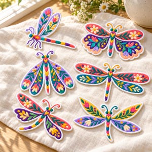 May include: A collection of six embroidered patches shaped like dragonflies and butterflies. Each patch features vibrant, multi-colored floral and leaf patterns in shades of pink, purple, yellow, green, and blue with white borders.