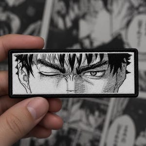 Puede incluir: Un parche rectangular bordado que presenta una ilustración en blanco y negro estilo manga de los ojos y cejas de un personaje. El diseño está encerrado en un borde negro grueso, mostrando un detallado trabajo de líneas y sombreado sobre fondo blanco.