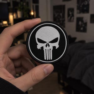May include: A circular embroidered patch featuring a white stylised skull design with sharp, angular eye sockets and vertical teeth lines set against a solid black background, enclosed by a thin white circular border and a black stitched edge.