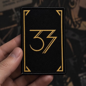 May include: A rectangular black patch featuring a gold embroidered border with corner accents and the number 37 in a stylised gold font centred within the frame. The design is stitched with a clean finish and a consistent gold thread colour.