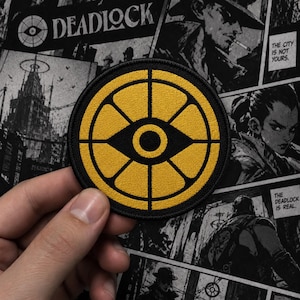Könnte beinhalten: Ein kreisförmiger bestickter Aufnäher mit einem goldgelben Raddesign und einem zentralen Augensymbol. Der Aufnäher hat einen dicken schwarzen Rand und schwarze Linien, die die gelben Segmente unterteilen. Das Design ist symmetrisch und grafisch.