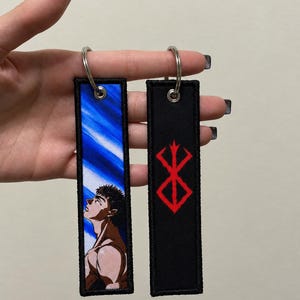 Pode incluir: Dois porta-chaves retangulares de tecido com argolas de metal. Um apresenta um fundo de céu azul com uma ilustração de perfil de uma personagem de cabelo escuro a olhar para cima. O outro é preto com um símbolo vermelho bordado.