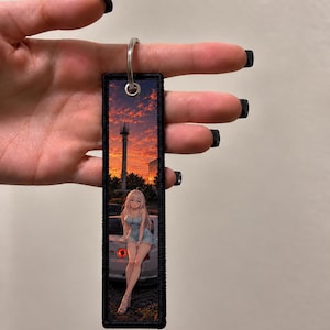 May include: A rectangular fabric keyring with a black embroidered border and a silver metal ring. The design features a sunset landscape with a tall communication tower and an anime-style character with long light hair sitting on a white car.