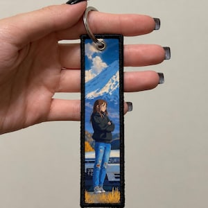 May include: A rectangular fabric keyring with a black stitched border featuring an anime-style illustration of a person in a black hoodie and blue jeans standing in front of a white car with a mountain landscape and blue sky in the background.