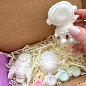 Op de afbeelding: Een wit keramieken astronautfiguurtje, een raket en een wereldbol in een kartonnen doos gevuld met papierversnippering. De set bevat kleine ronde verfpotjes en een houten penseel. De voorwerpen zijn ongeverfd en hebben een matte afwerking.