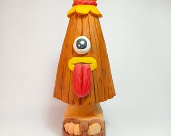 May include: A hand-carved wooden figurine featuring a tall, conical brown body with a single large eye, a yellow mouth, and a long red tongue. A small yellow head sits on top, and the base shows a foot wearing a brown sandal with visible wood grain.