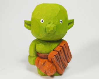 May include: A small, hand-carved wooden figurine of a green creature with large ears, a rounded head, and a prominent nose. The figure has white eyes with black pupils and is holding a textured, orange-colored object across its midsection.