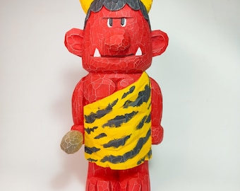 May include: A hand-carved wooden figurine of a red oni demon with dark gray hair, yellow horns, and white fangs. The figure wears a yellow loincloth with black tiger stripes and holds a small, textured, light brown stone-like object in one hand.