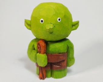 May include: A small, hand-carved wooden figurine of a green creature with a large, rounded head, pointed ears, and a small nose. The figure has simple black and white eyes and is wearing a brown garment while holding a brown staff in its hand.