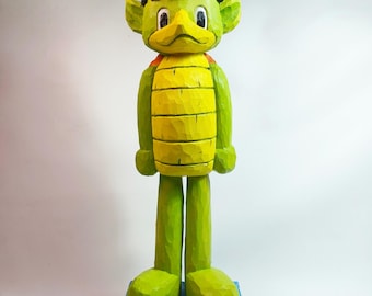 May include: A hand-carved wooden figurine of a green creature with a yellow segmented torso, black hair, and large white eyes. The figure stands on a textured blue rectangular base, featuring visible wood grain and carved details throughout the body.