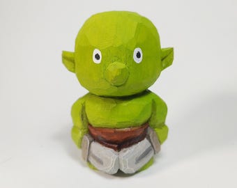 May include: A small, hand-carved wooden figurine of a green creature with large ears, a rounded head, and a pointed nose. The figure features white eyes with black pupils, a brown waist section, and light gray leg coverings with visible carved textures.