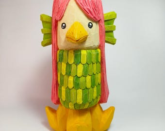 May include: A hand-carved wooden figurine featuring a creature with bright pink hair, a yellow beak, and black eyes. The body is covered in overlapping green and light green scales, with green fins on the sides and yellow feet at the base.