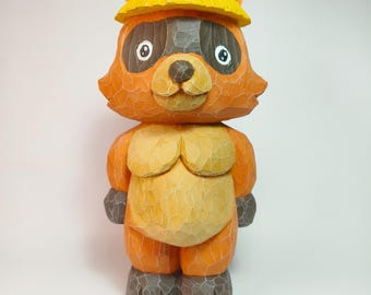 May include: A hand-carved wooden figurine of a raccoon wearing a yellow conical hat. The raccoon has orange fur, dark gray eye patches, a light tan muzzle, and dark gray feet. The surface shows visible wood grain texture throughout the carving.