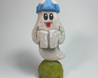 May include: A hand-carved wooden figurine of a white ghost with a blue headband, black eyes, and a red tongue sticking out. The ghost is positioned on a textured, rounded green base, showcasing visible wood grain and carved chisel marks throughout.