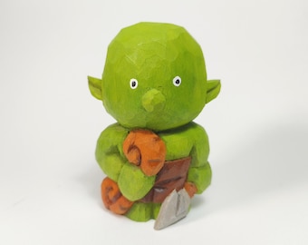 May include: A small, hand-carved wooden figurine of a green creature with large ears, holding a round orange object in one hand and a grey tool in the other. The figure has a smooth, rounded head, a brown waist wrap, and visible wood grain texture.