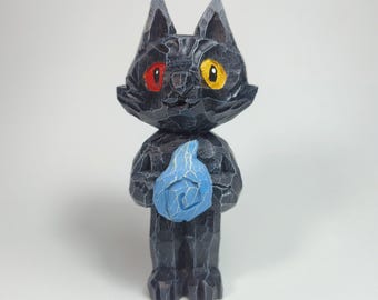 May include: A hand-carved dark charcoal cat figurine with a faceted texture. The cat has one red eye and one yellow eye, and it holds a light blue flame-shaped object in its front paws. The figure has pointed ears and a stylized, angular design.