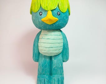 May include: A hand-carved wooden figurine of a creature with a teal body, a bright yellow beak, and a vibrant lime green bowl-cut hairstyle. The surface features visible wood grain textures, black circular eyes, and a rounded, segmented belly.