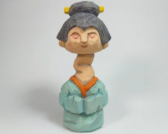 May include: A hand-carved wooden figurine featuring a stylized figure with a long neck, dark grey hair styled in a bun with yellow accents, and a light blue robe with an orange collar. The figure has closed eyes and a neutral facial expression.