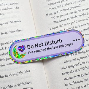 May include: A rectangular bookmark with rounded corners featuring a light purple background and a holographic border. The text reads Do Not Disturb and I've reached the last 100 pages, accompanied by a small holographic heart and crescent moon icon.