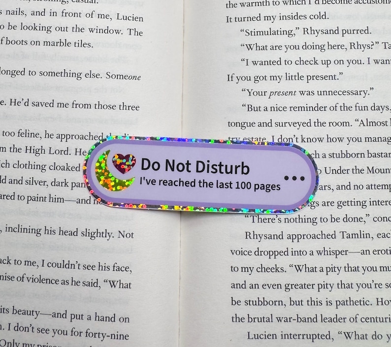 May include: A rectangular bookmark with rounded edges featuring a light purple background, a holographic crescent moon and heart icon, and the black text "Do Not Disturb" followed by "I've reached the last 100 pages" and three dots.