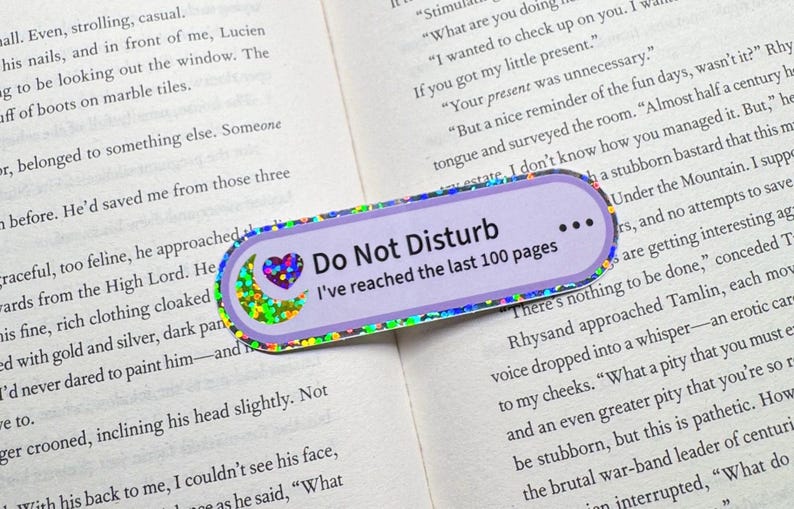 May include: A rectangular bookmark with rounded corners featuring a light purple background and a holographic border. The text reads Do Not Disturb and I've reached the last 100 pages, accompanied by a small holographic heart and crescent moon icon.