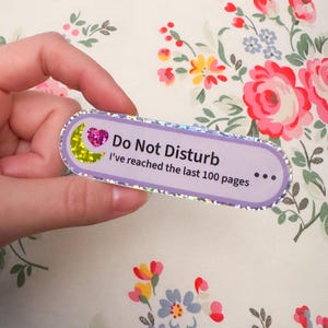 May include: A rectangular sticker with rounded edges and a holographic silver border. The sticker is light purple and features a yellow glitter crescent moon, a pink glitter heart, and the text: Do Not Disturb I've reached the last 100 pages.