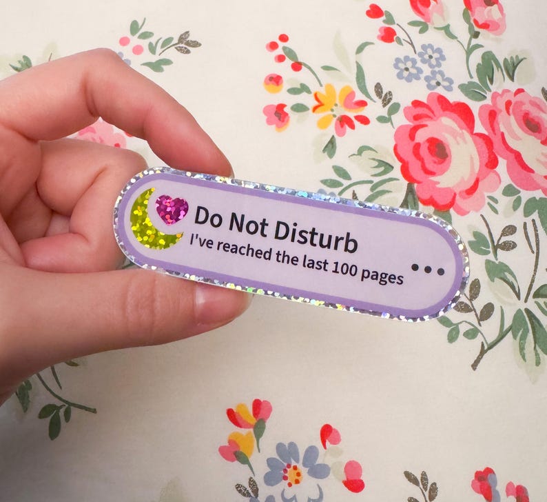 May include: A rectangular sticker with rounded edges and a holographic silver border. The sticker is light purple and features a yellow glitter crescent moon, a pink glitter heart, and the text: Do Not Disturb I've reached the last 100 pages.