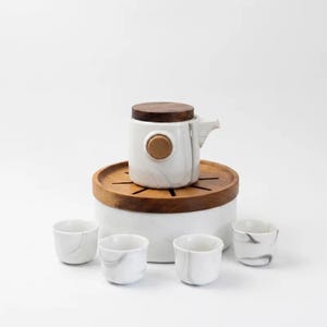 Könnte beinhalten: Ein weißes Keramik-Teeservice bestehend aus einer Teekanne mit dunklem Holzdeckel und rundem Holzknopf, platziert auf einem runden weißen Sockel mit geschlitztem Holztablett. Vier kleine weiße Keramiktassen mit grauem Marmormuster.