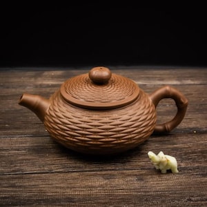 Peut inclure: Une théière en argile brune avec un motif tressé texturé sur le corps et le couvercle, dotée d'un bec verseur incurvé et d'une anse en forme de tige de bambou, accompagnée d'une petite figurine d'éléphant en pierre sculptée claire.