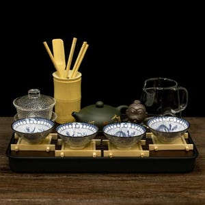 May include: A tea set featuring four white ceramic bowls with blue dragon patterns, each resting on a small wooden stand. The set includes a dark green teapot, a clear glass pitcher, a textured glass lidded bowl, and a bamboo utensil holder.