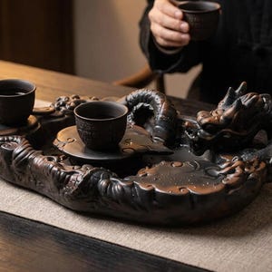 Pode incluir: Uma bandeja de chá de cerâmica castanho-escura com um motivo de dragão esculpido, escamas detalhadas e uma cabeça elevada. A bandeja inclui duas pequenas chávenas de chá a condizer em cerâmica castanho-escura com padrões texturados.