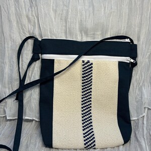 May include: A crossbody bag featuring a rectangular shape with a black canvas exterior and a central vertical panel of cream-colored fabric. The bag includes a white zippered closure at the top and a thin black shoulder strap attached to the sides.