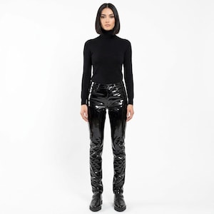 Puede incluir: Un suéter negro de cuello alto y manga larga combinado con pantalones de cuero sintético negro de alto brillo. Los pantalones cuentan con cierre de botón y cremallera, trabillas para cinturón y un acabado reflectante que resalta la textura.