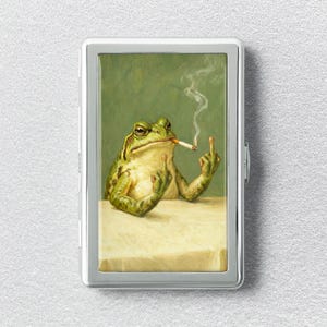 May include: A rectangular silver metal cigarette case featuring a printed illustration of a green frog sitting at a table, smoking a cigarette, and gesturing with its middle fingers against a muted green background.