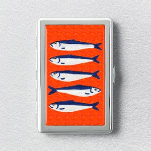 May include: A rectangular metal cigarette case featuring a vibrant orange background with a repeating pattern of five horizontal blue and white fish stacked vertically. The case has a polished silver-toned frame and a hinged opening mechanism.