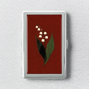 May include: A rectangular metal cigarette case with a polished silver frame and a deep red center panel. The center features a botanical illustration of a lily of the valley plant with white bell-shaped flowers and two large green leaves.