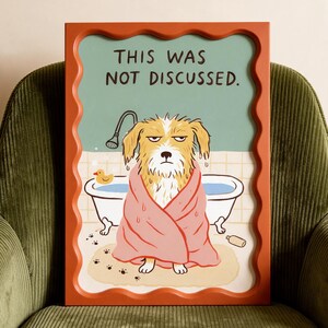 May include: A rectangular wall art print in a wavy orange frame featuring a cartoon illustration of a wet, grumpy dog wrapped in a pink towel in a bathroom. The text "THIS WAS NOT DISCUSSED." is printed in black at the top above the bathtub.