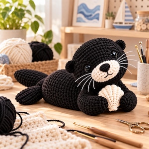 May include: A black crocheted otter plush toy with a cream-colored muzzle, black safety eyes, and long white whiskers. The otter is holding a small, cream-colored crocheted seashell in its front paws while lying on a wooden surface.