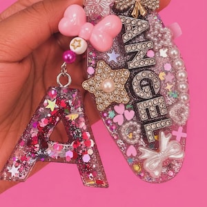 May include: A resin keychain set featuring an oval plaque with the word ANGEL in rhinestone letters, decorated with pearls, bows, and stars. A separate letter A charm is attached, both filled with pink and silver glitter and confetti.