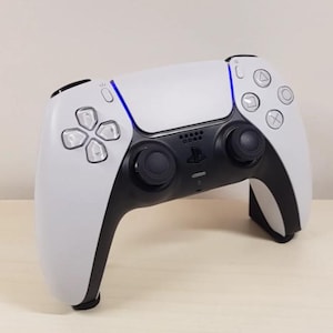 Pode incluir: Um comando de videojogos sem fios branco e preto com um painel tátil central com faixas de luz azul, dois joysticks pretos, um pad direcional e quatro botões de ação com símbolos geométricos incluindo triângulo, círculo, cruz e quadrado.