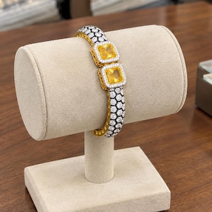 May include: A hinged bangle bracelet featuring two prominent square-cut yellow stones set in silver-toned frames. The band is decorated with a dense pattern of small, round, clear stones set against a dark, honeycomb-textured background.