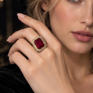 Puede incluir: Un anillo de oro con una gran piedra preciosa rectangular de color rojo intenso en el centro. La piedra está rodeada por un doble halo de pequeñas piedras claras y brillantes engastadas en una banda de oro, creando una joya detallada.