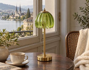Peut inclure: Une lampe de table dotée d'un abat-jour en verre vert lime translucide et nervuré en forme de pétale de fleur, soutenu par une tige métallique fine en or brossé et une base circulaire, posée sur une surface en bois sombre.