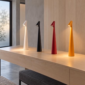 May include: Four tall, slender, conical table lamps arranged in a row. The lamps are white, black, red, and yellow, each featuring a small, angled light source at the top that casts a warm glow downward onto the surface below the lamp base.