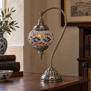 May include: A table lamp featuring a rounded glass shade decorated with a colorful mosaic pattern of small diamond-shaped tiles in red, blue, green, and yellow. The lamp has a curved, twisted metal stand with an antique silver finish.