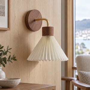 Könnte beinhalten: Eine Wandleuchte mit einem runden Sockel aus dunklem Holz, einem geschwungenen Messingarm und einem oberen Verbindungsstück aus dunklem Holz. Die Lampe hat einen weißen, kegelförmigen, plissierten Stoffschirm, der nach unten gerichtet ist.